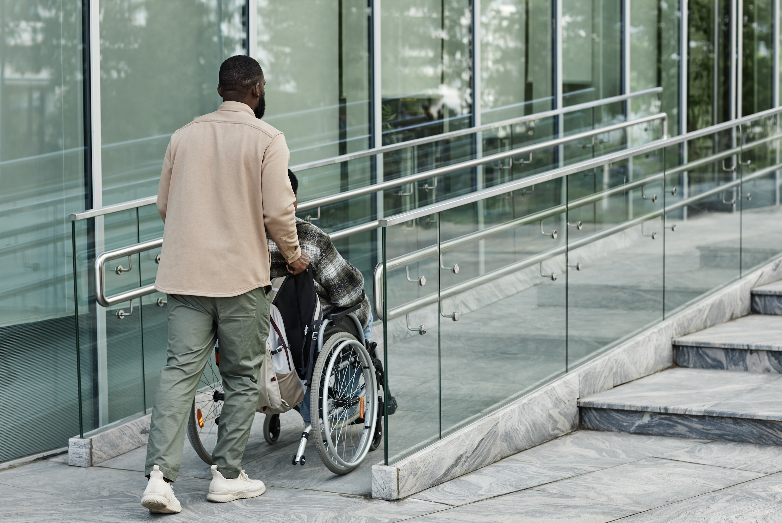 Man Walking Person in Wheelchair Up A Ramp to Access A Building