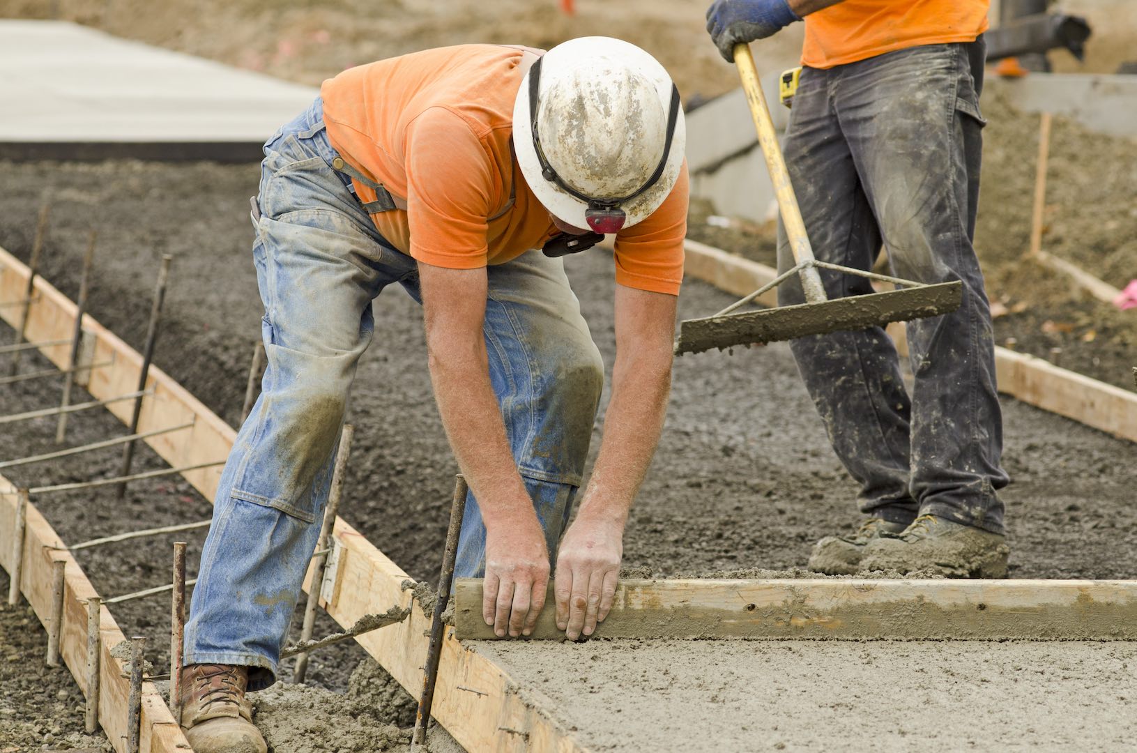 Construction workers pour and level concrete within a wooden form, preparing a sidewalk or pathway as part of an infrastructure project. One worker smooths the surface while another ensures proper alignment.