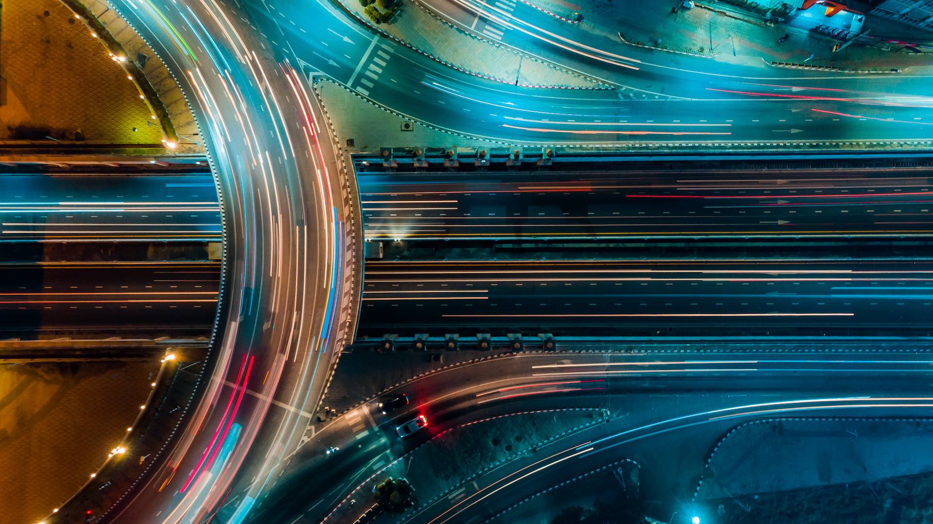 Aerial view of a brightly lit highway interchange at night, with colorful light trails from moving vehicles creating arcs and lines across multiple overpasses and roadways.