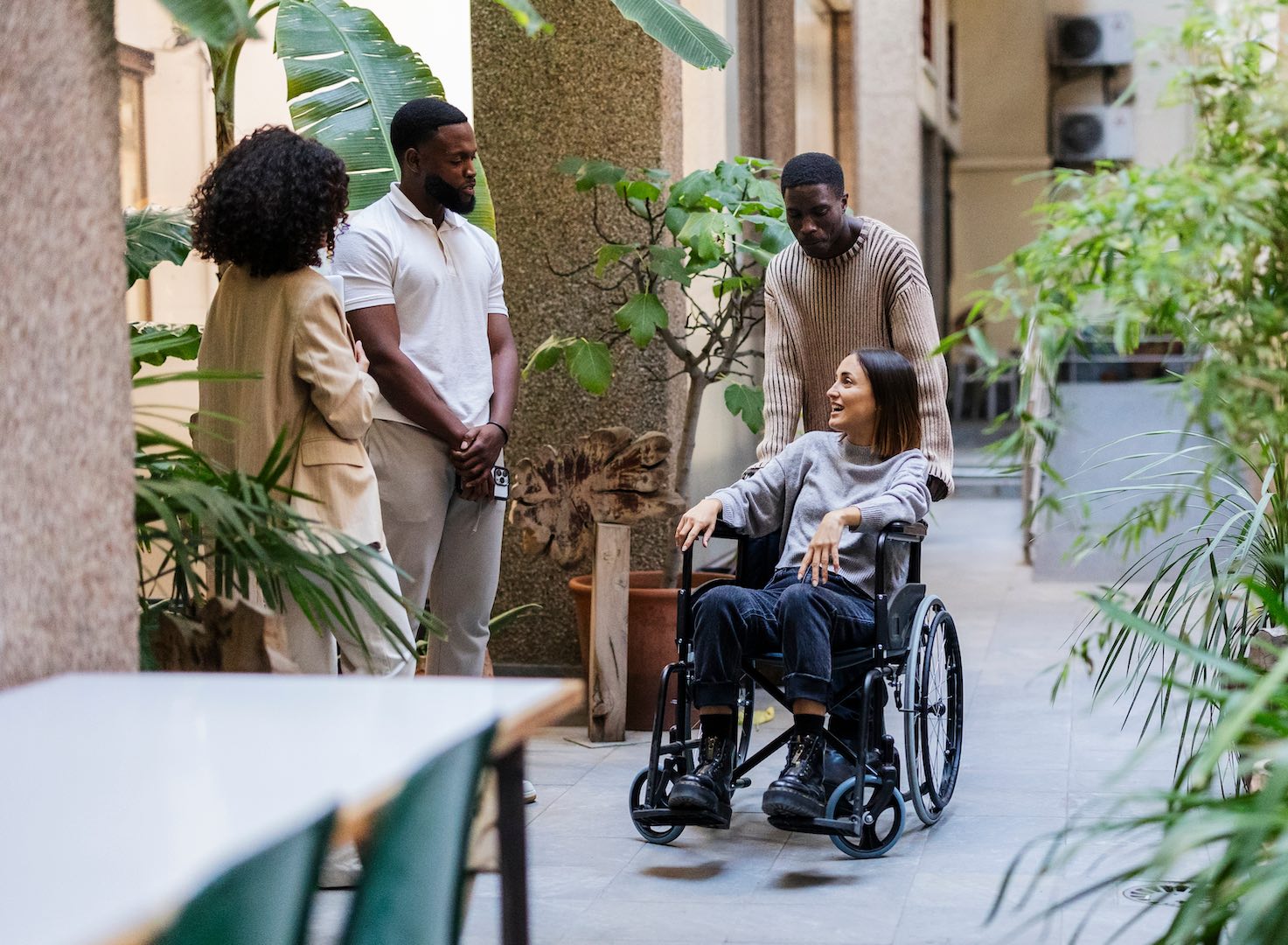 A young woman in a wheelchair smiling and talking with three friends in a modern, plant-filled outdoor corridor, illustrating inclusive and accessible social spaces.