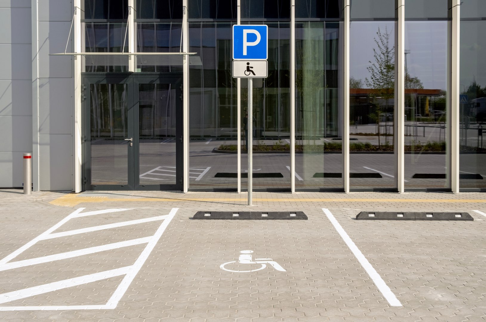 An accessible parking space marked with a wheelchair symbol is situated in front of a modern glass office building, with a designated access aisle and signage indicating reserved parking for people with disabilities.