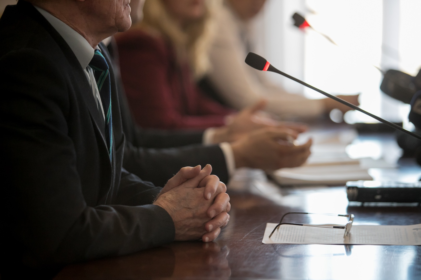 A panel of officials sits at a conference table with microphones, folded hands, and documents in front of them, suggesting a formal discussion or public meeting.